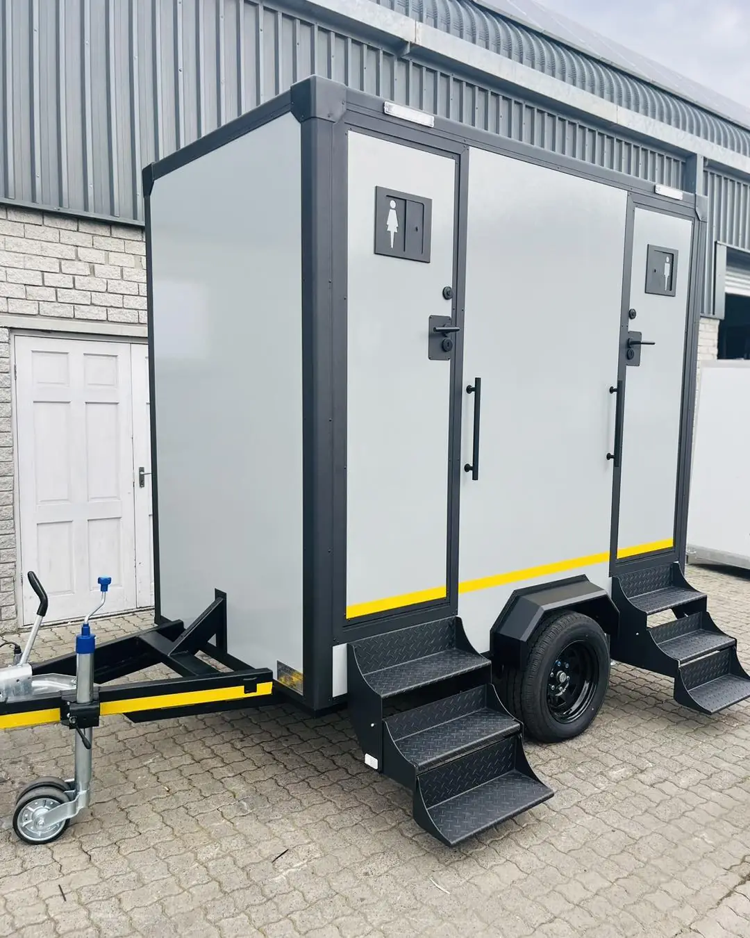 Gray two-door restroom trailer with fold-down metal steps parked on pavers